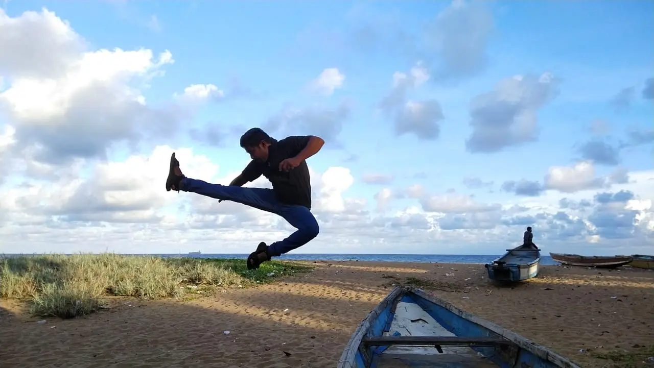 A Gladiators student mid jump-kick on the beach, fishing boats behind him