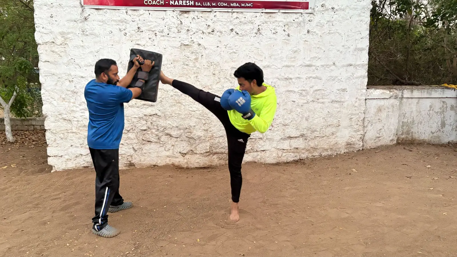 A coach holds a kick shield as a student throws a roundhouse kick against the club's whitewashed wall