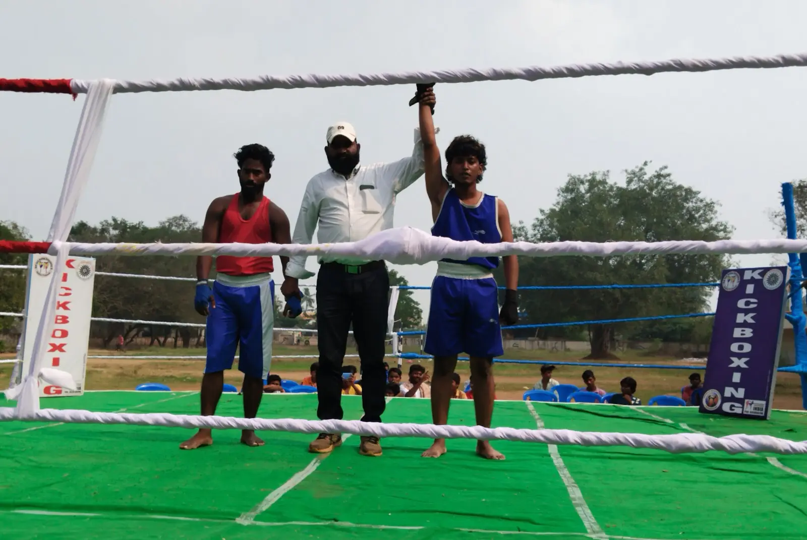 Two young fighters in red and blue with the referee in an outdoor kickboxing ring