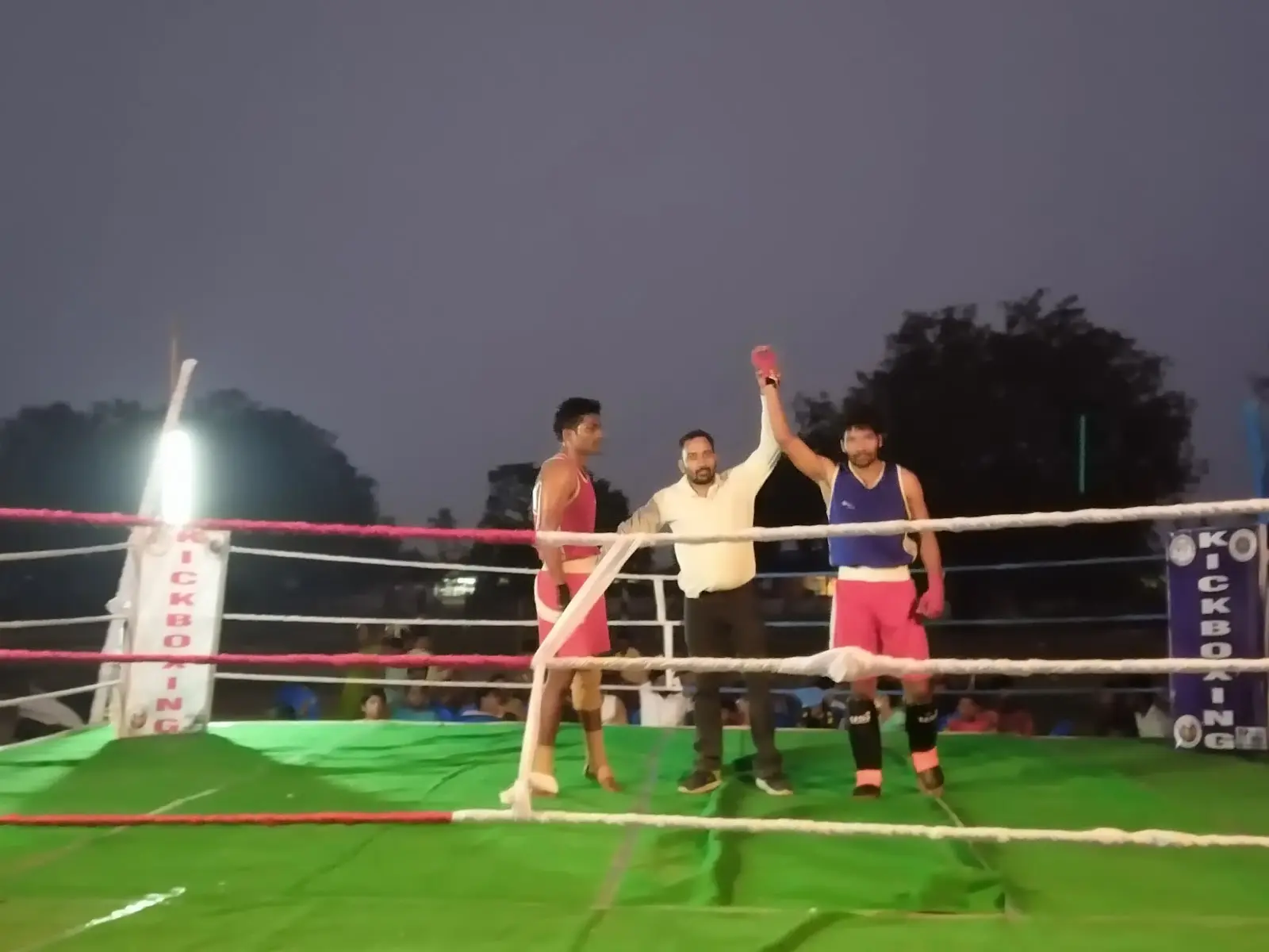 Referee raises a fighter's hand in an outdoor kickboxing ring at night